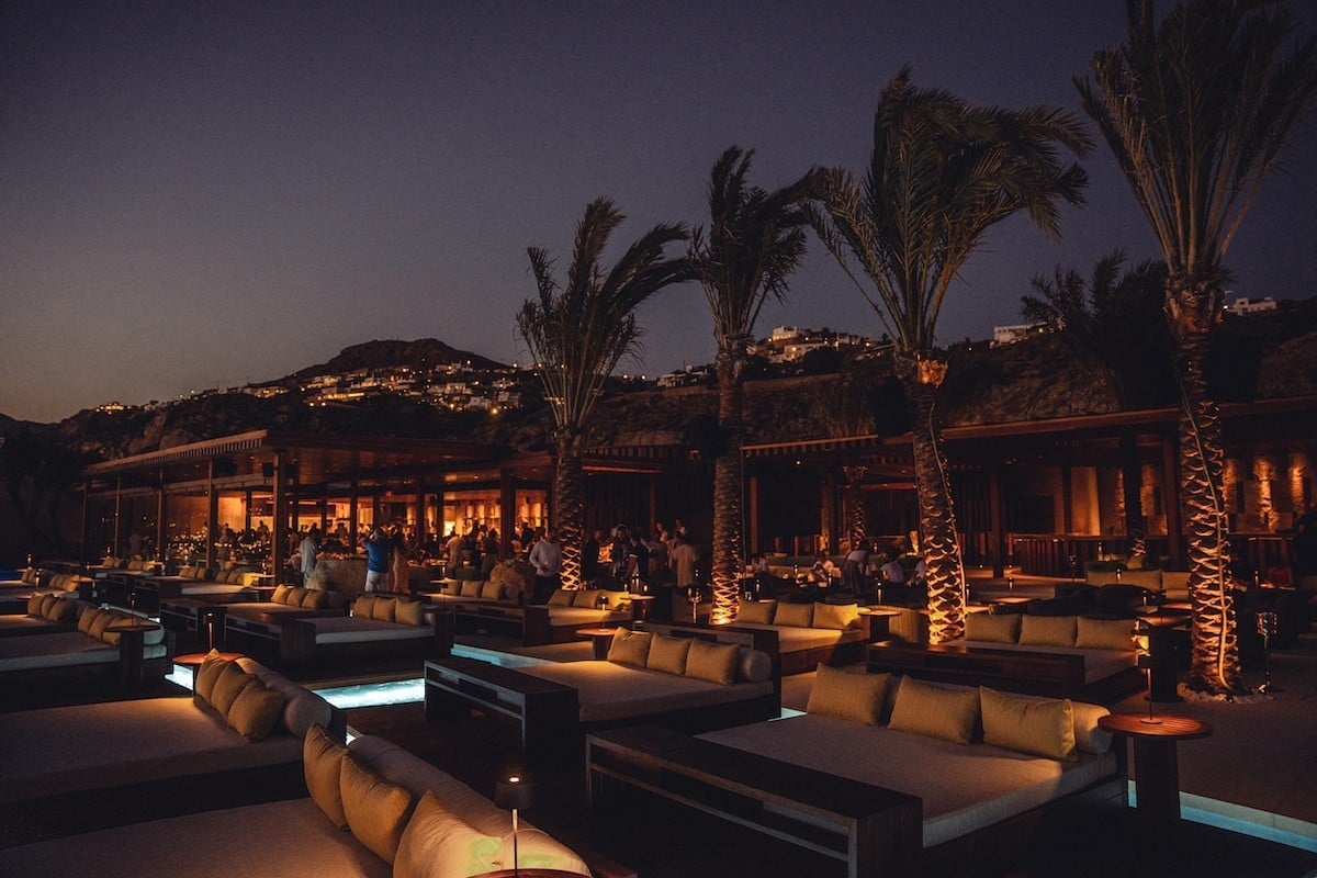 Outdoor lounge area at dusk with illuminated palm trees, seating areas, and buildings on a hillside at night