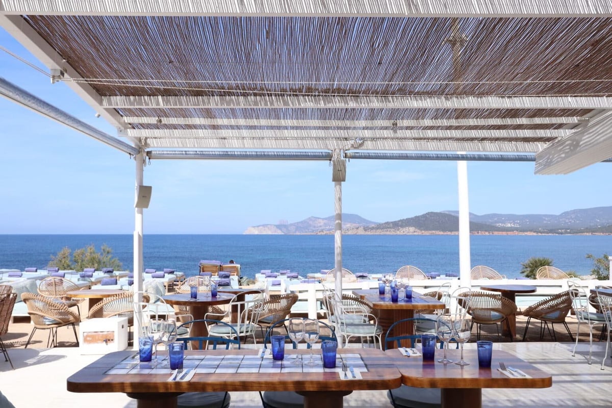Beachfront restaurant terrace with pergola overhead, wooden furniture, and panoramic blue sea and mountains in background
