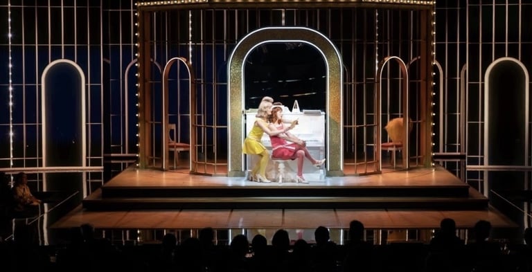 Theater stage with ornate archway set featuring performers in colorful costumes under dramatic lighting with audience silhouettes in foreground