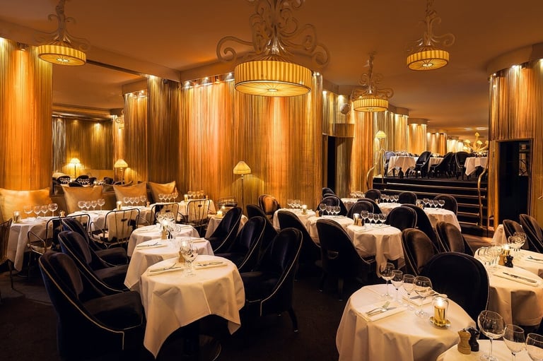 Elegant fine dining restaurant interior with round tables dressed in white linens, black velvet chairs, and ornate golden pendant lighting fixtures