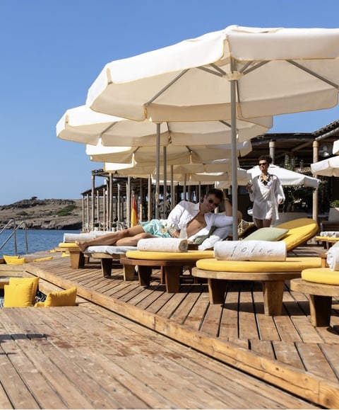 Beachside lounge with wooden deck, cream umbrellas, yellow cushioned daybeds, and blue water view