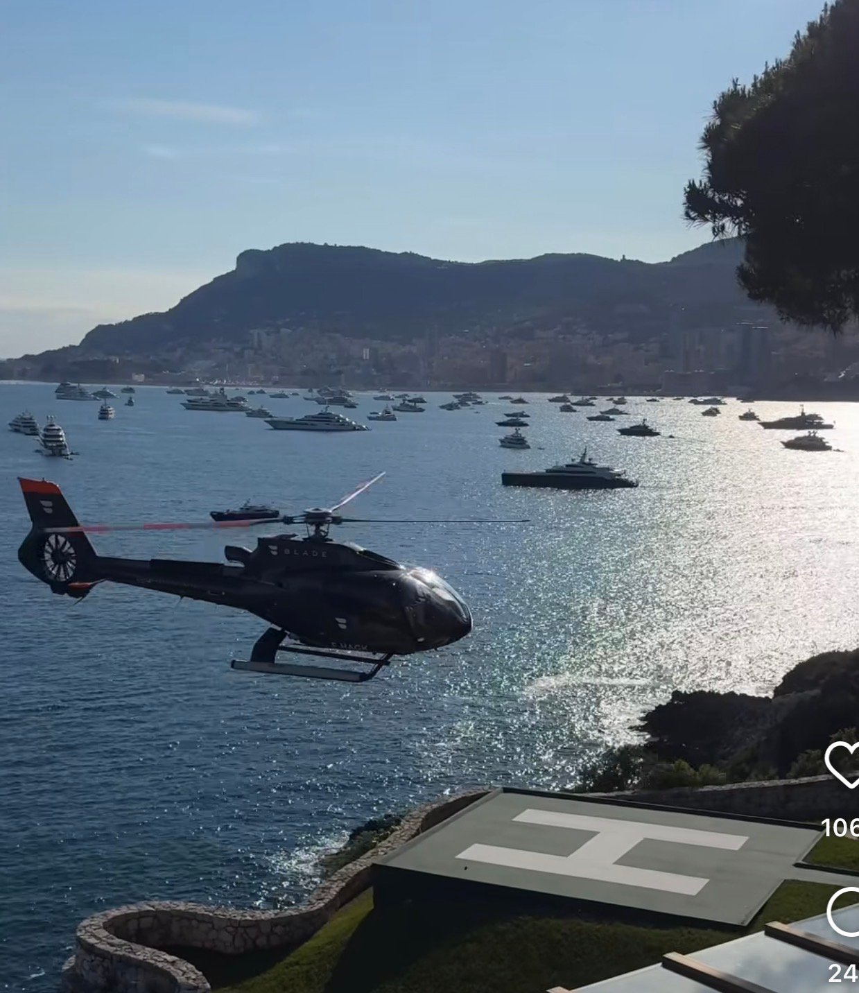 Black helicopter hovering over turquoise water near rocky coastline with moored boats and mountains in background