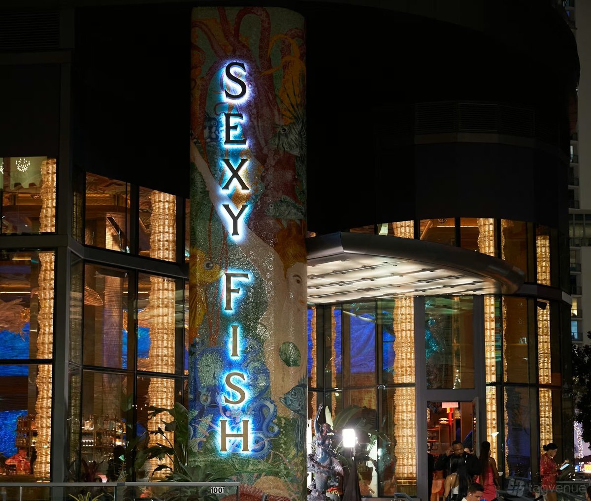 Neon sign reading SEXY FISH illuminated in blue on a building facade at night with brick and glass storefronts
