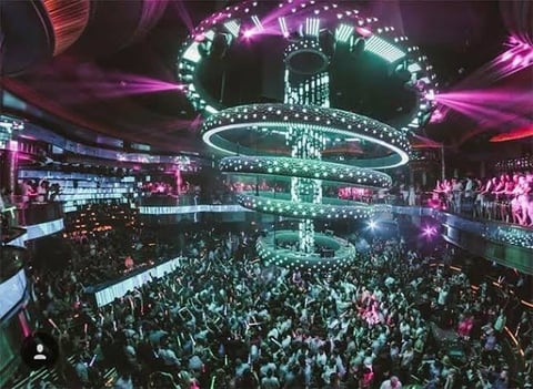 Nightclub interior with packed dance floor, neon purple and green lighting, and elaborate glowing chandelier-style fixture overhead