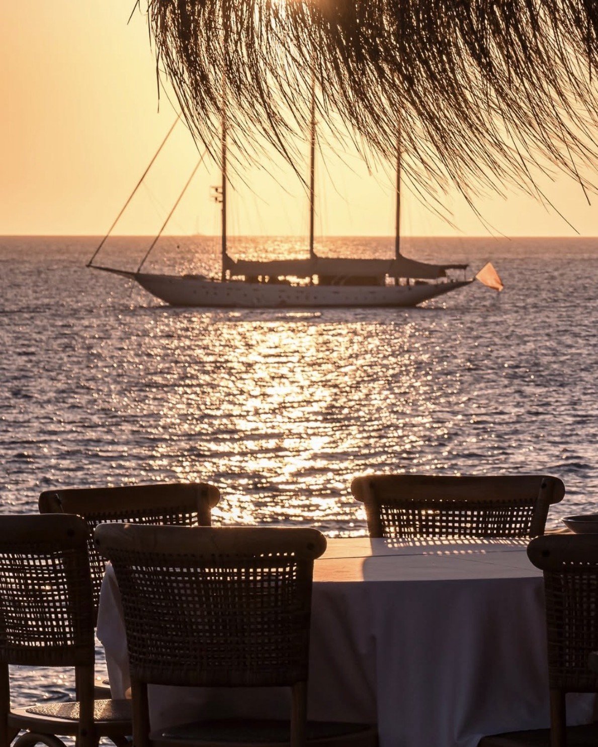 Silhouetted sailboat anchored at sunset with woven chairs and table in foreground, viewed from beachfront restaurant or terrace