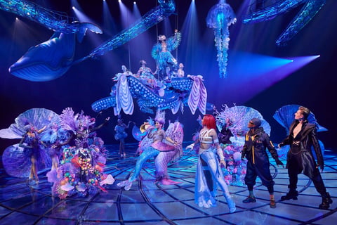 Elaborate stage performance with performers in fantastical costumes under dramatic blue lighting and suspended ethereal structures