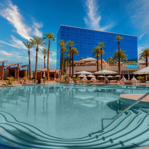 Luxury resort pool with palm trees and modern blue glass building under clear blue sky with white umbrellas and loungers