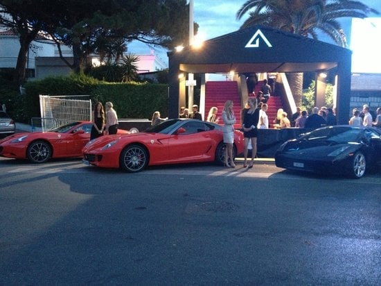 Red and dark blue sports cars displayed outside an illuminated storefront at dusk with visitors nearby