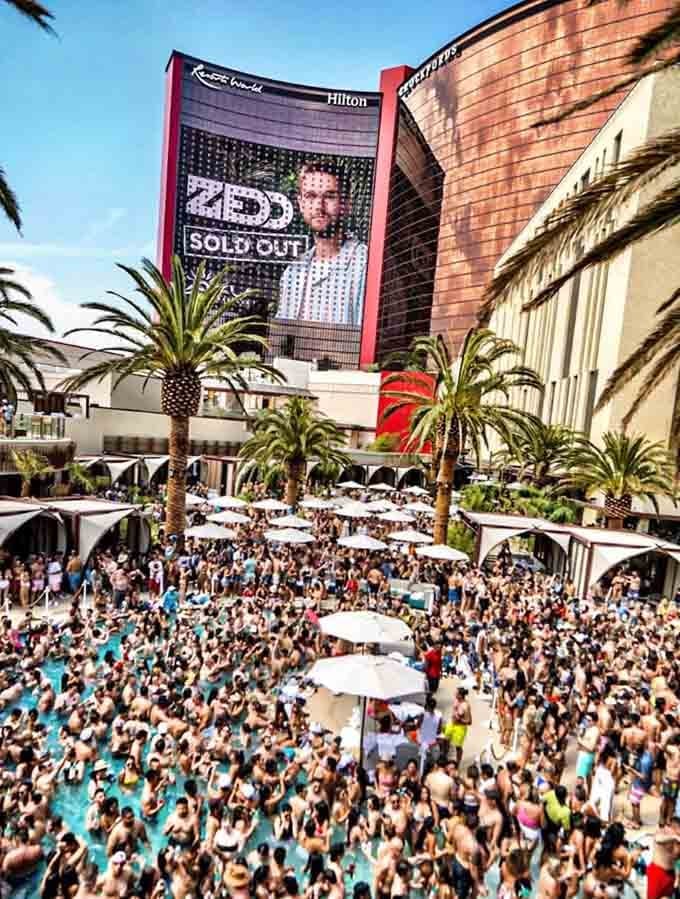 Crowded outdoor pool party at Hilton hotel with large billboard showing sold out concert, palm trees, and hundreds of people gathered in swimwear