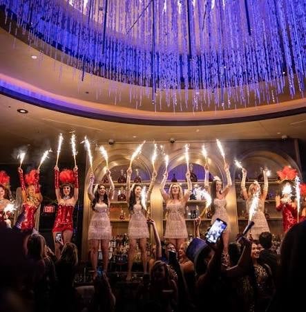 Women in white dresses performing on stage with dramatic blue lighting and a crowd below at an indoor concert venue