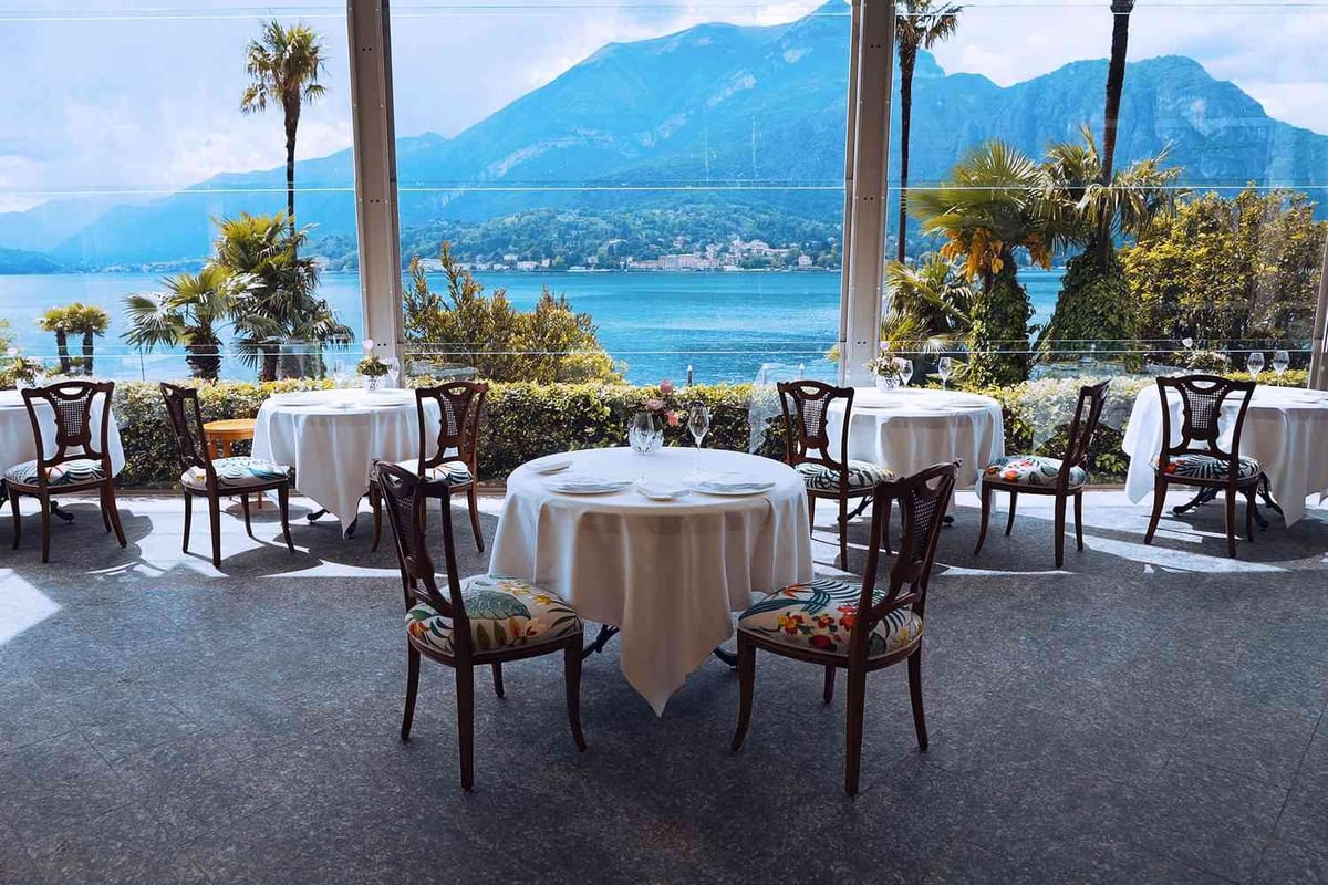 Elegant lakeside restaurant terrace with round tables, white tablecloths, and mountain views across blue water