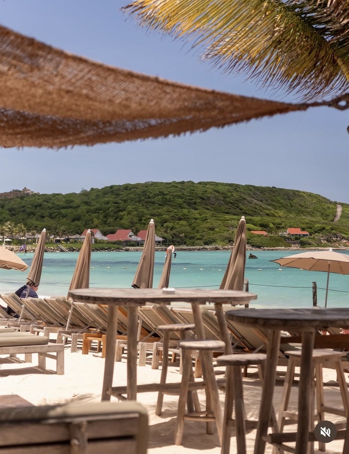 Wooden beach chairs and umbrellas on white sand facing turquoise water with green hillside and palm trees