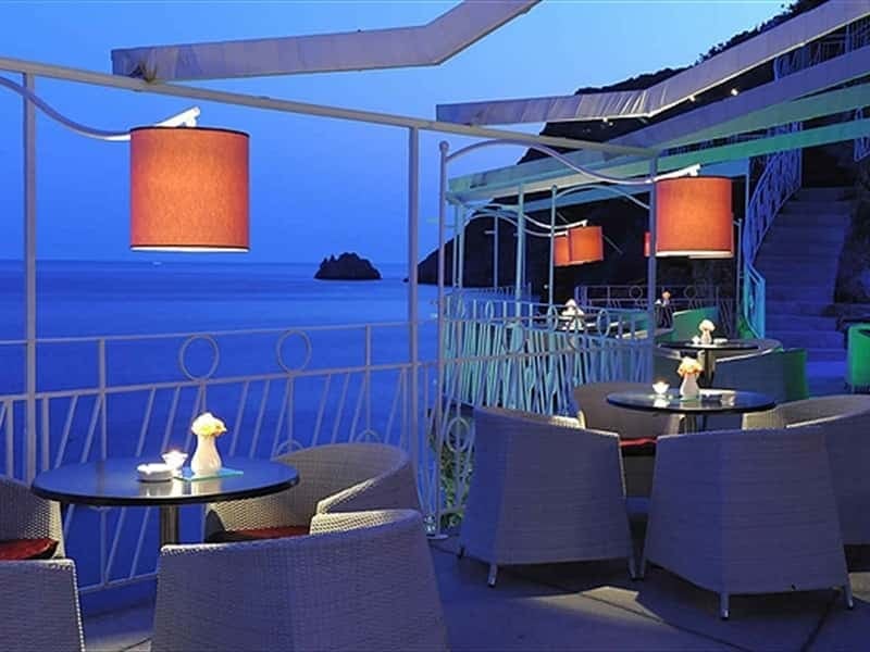 Modern rooftop lounge at dusk with blue lighting, elegant furniture, and orange hanging lamps overlooking the sea
