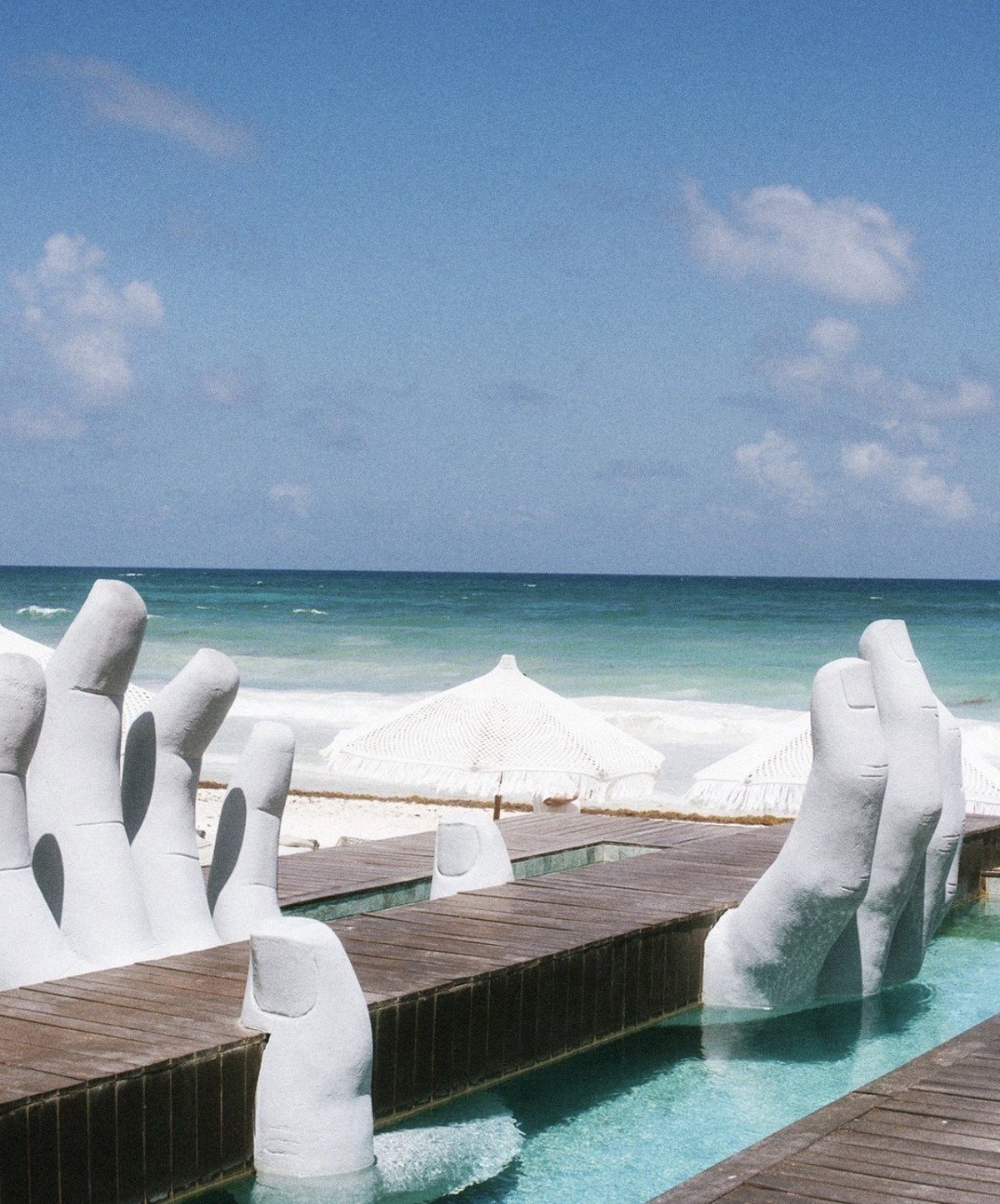 Tropical beach venue with white sculptural hand installations flanking a wooden dock and white tent overlooking turquoise water and white sand beach