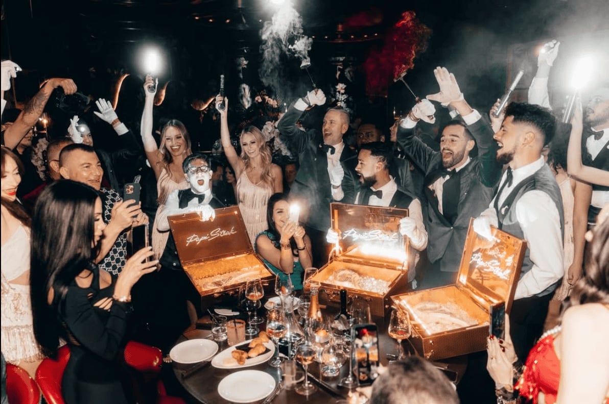 Group of people celebrating at a formal event with gold gift boxes on table, confetti falling, and guests with raised hands cheering