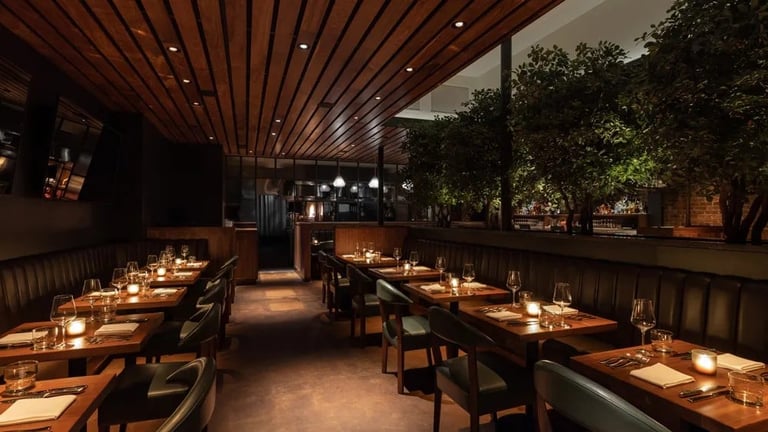 Elegant restaurant interior with long wooden tables, place settings, candlelit ambiance, and lush green plants along the windows