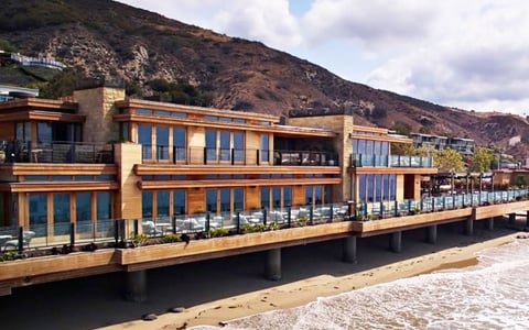Modern waterfront building with blue windows and wooden facade built on stilts over water, with desert hills in background