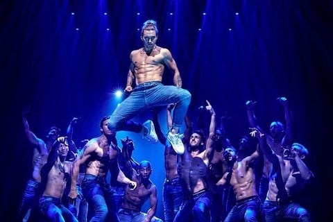 Performer on stage under blue lighting, viewed from behind with arms on hips in a confident pose