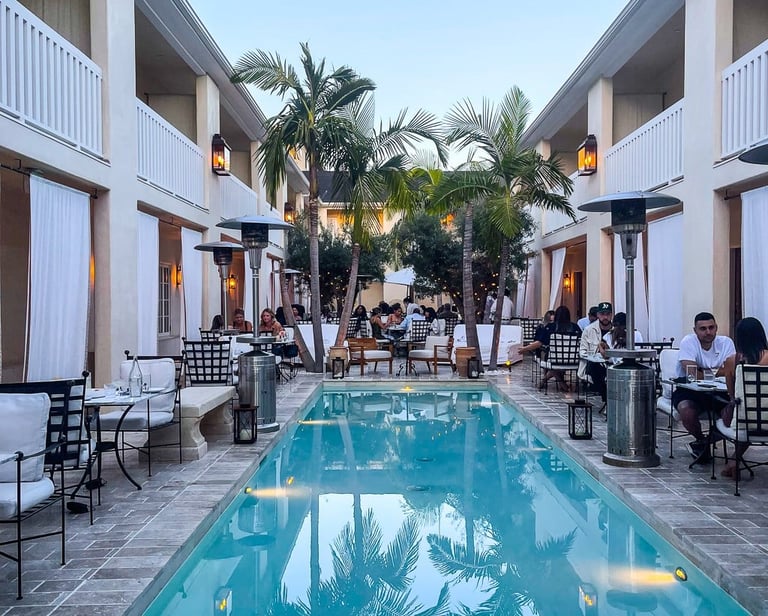 Elegant courtyard with a long rectangular pool flanked by white buildings, palm trees, and outdoor seating areas at dusk