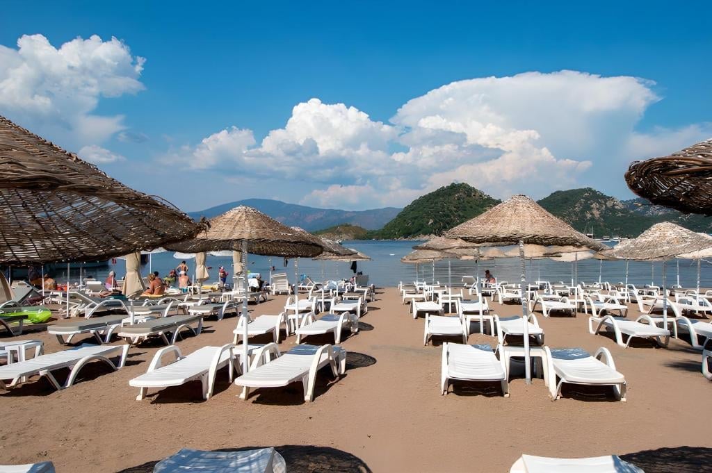 Beach with rows of white lounge chairs, thatched umbrellas, and mountains under blue sky with clouds