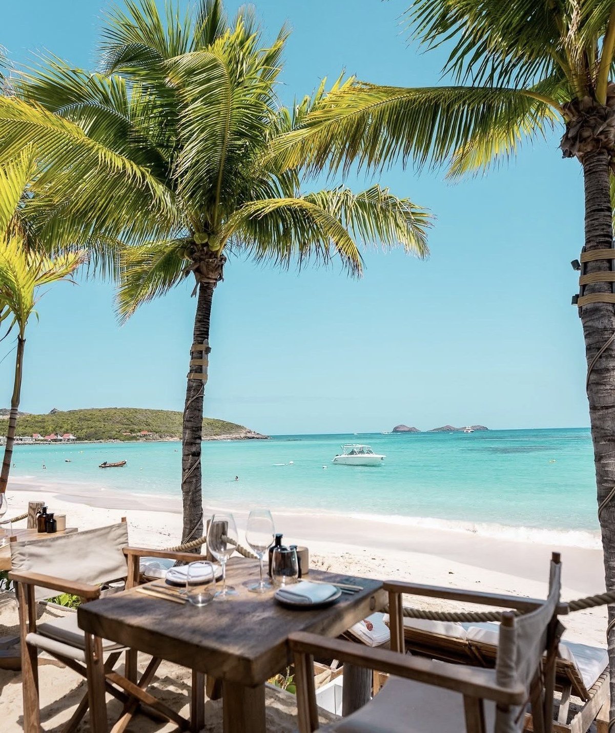 Outdoor dining setup on tropical beach with palm trees, white sand, turquoise water, and boats
