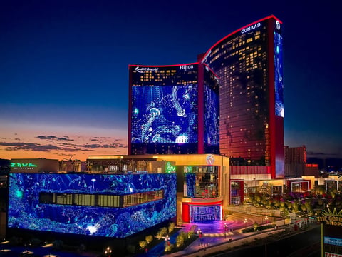 Illuminated resort casino at dusk with red and blue LED displays, glowing architectural elements, and surrounding cityscape at twilight