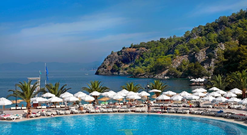Luxury resort with turquoise pool, white umbrellas, palm trees, and forested hillside overlooking a blue coastal bay