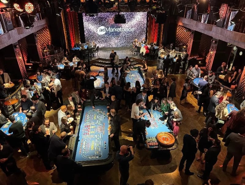 Overhead view of a casino floor with illuminated gaming tables, crowds of people, and red stage lighting