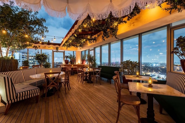 Elegant rooftop restaurant with wooden deck, striped seating, hanging greenery, and panoramic city views at dusk