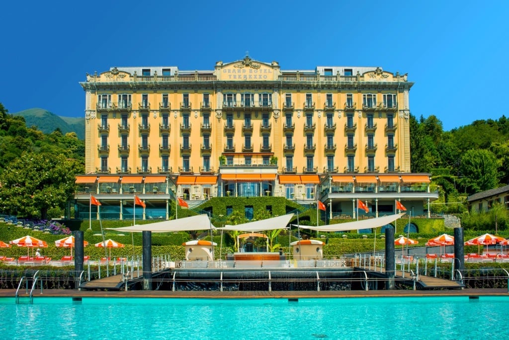 Grand yellow waterfront hotel with terrace on a bright day, turquoise lake water in foreground, surrounded by lush green mountains