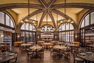 Elegant restaurant dining room with ornate wooden ceiling, large arched windows, and multiple round tables set for dining