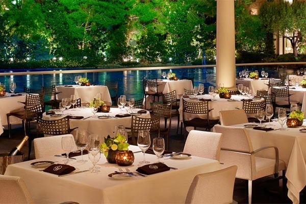 Upscale outdoor dining venue with round tables, cream tablecloths, patterned chairs, and a water view backdrop lined with lush greenery