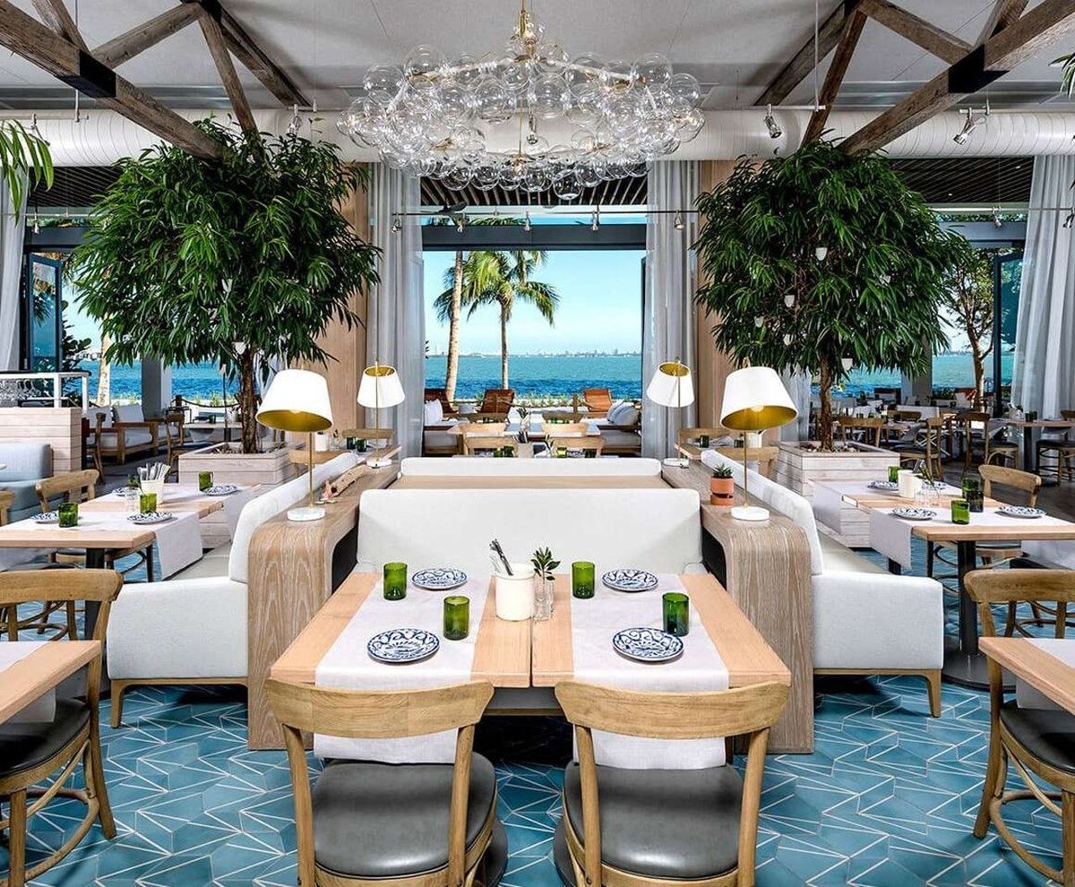 Luxury coastal restaurant interior with ocean view, elegant chandelier, wooden dining tables, white seating, and tropical plants.