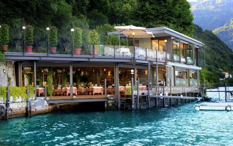 Modern waterfront restaurant built on a dock over turquoise lake water, surrounded by forested mountains