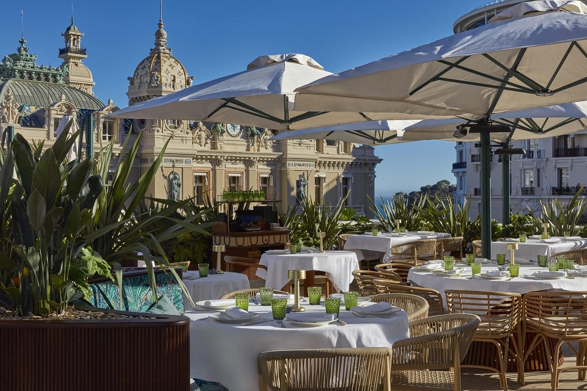 Elegant outdoor rooftop terrace with white table settings, wicker chairs, beige umbrellas, and potted plants overlooking ornate European buildings under blue sky