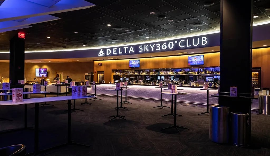 Interior of Delta Sky360 Club with illuminated signage, bar counter, blue lighting, and queue barriers in an airport lounge setting