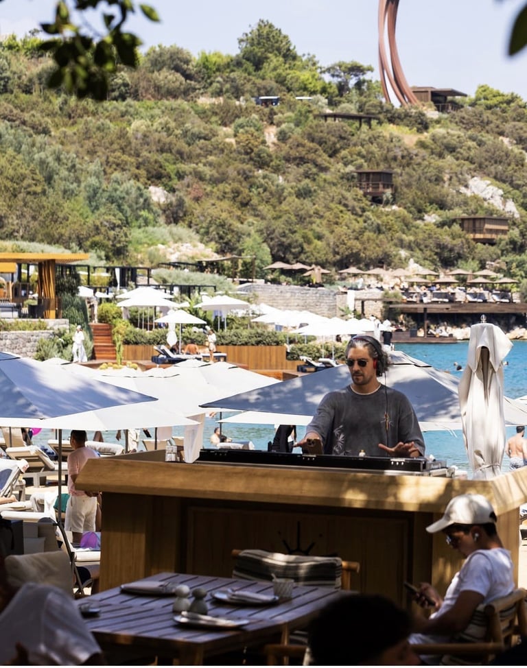 DJ performing at outdoor beach club with pool, hillside structures, and Mediterranean landscape in background