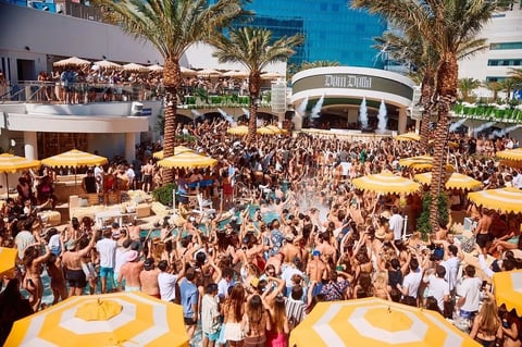 Aerial view of a crowded outdoor festival or event with yellow umbrellas, palm trees, and multiple levels of attendees in a modern plaza