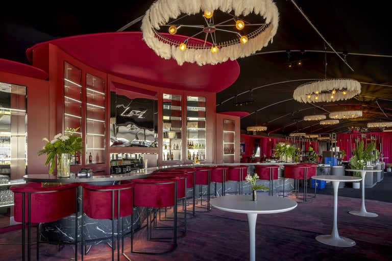 Modern restaurant interior with burgundy walls, illuminated bar shelving, hanging crystal chandeliers, and round white tables on polished floor.