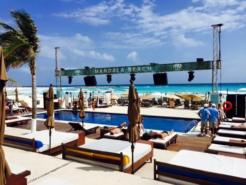 Beach club with blue pool, lounge chairs, stage setup, palm trees, and turquoise ocean under blue sky
