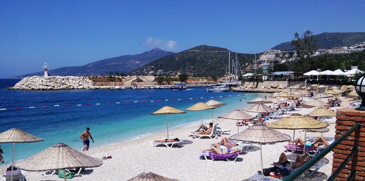 Scenic Mediterranean beach with turquoise water, white sand, beach umbrellas, sunbathers, sailing boats, and mountains in the background