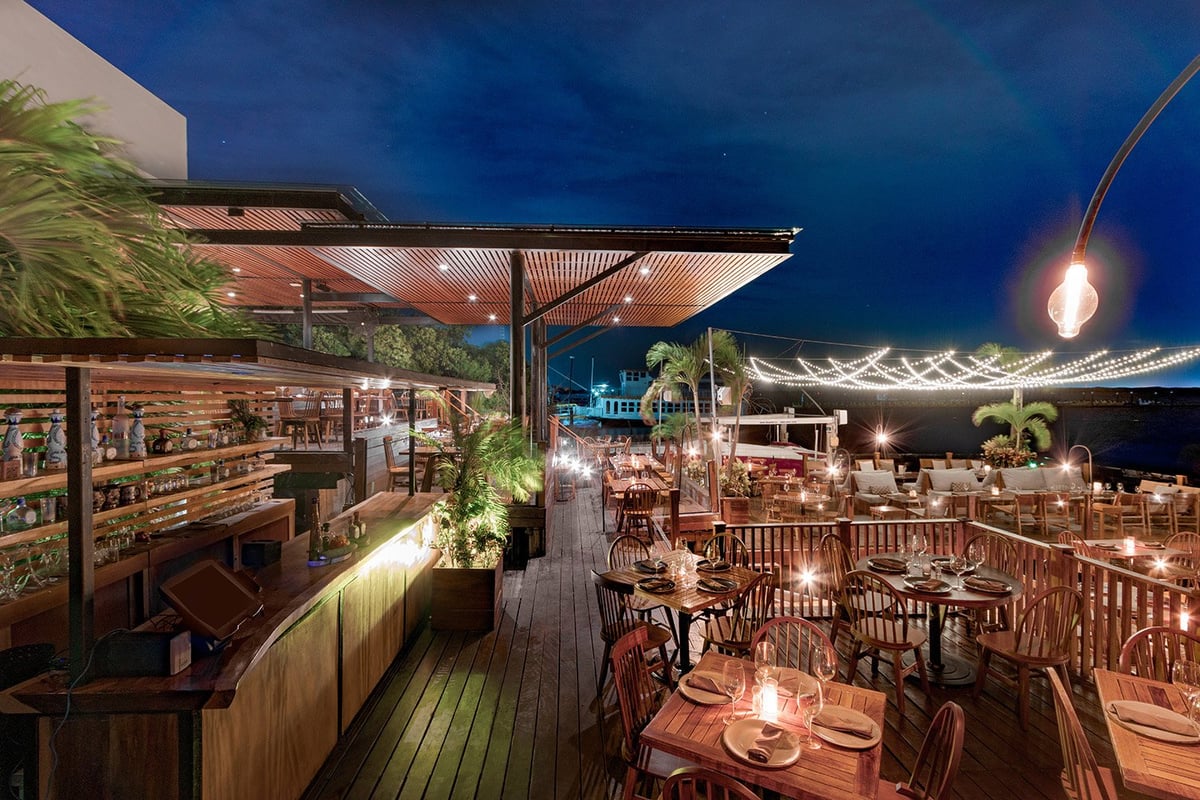 Upscale rooftop dining area at night with wooden deck, set tables, bar counter, pendant lighting, and city skyline in background