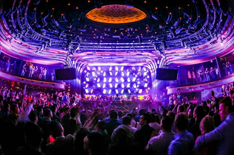 Crowded electronic music concert with purple and pink stage lighting, large video screens, and illuminated ceiling in a packed venue