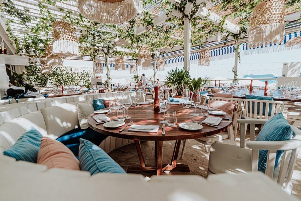 Elegant rooftop dining area with round wooden table set for dinner, white seating with blue pillows, ivy-covered walls, and Mediterranean architecture