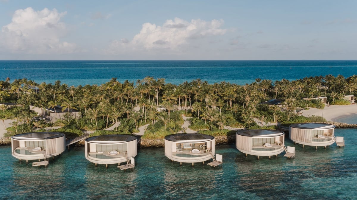 Row of modern overwater bungalows with thatched roofs along turquoise ocean near tropical island with palm trees