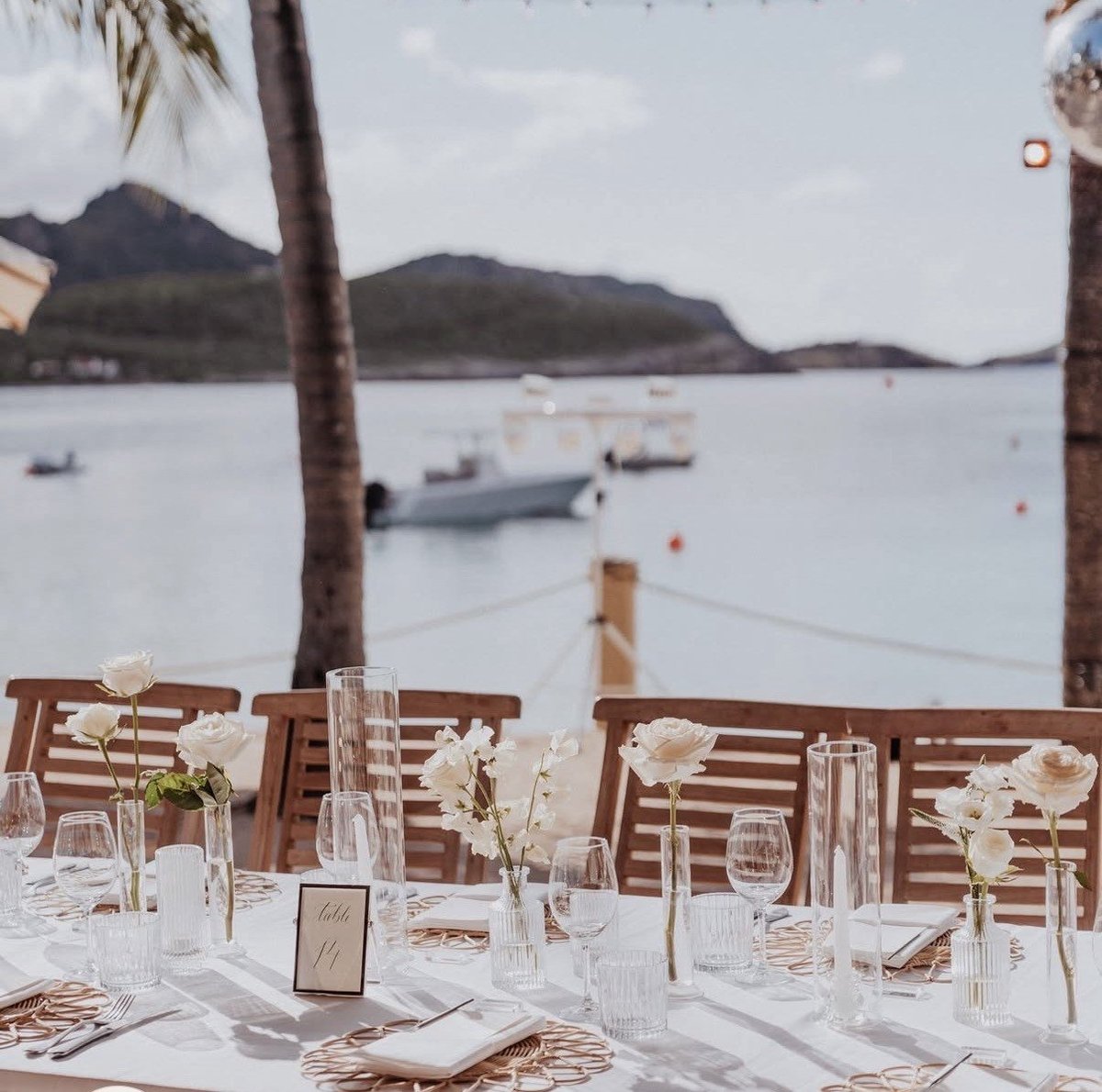 Elegant outdoor wedding reception table overlooking tropical bay with moored boats and palm trees
