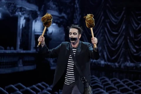 Man in dark theatrical costume with pale makeup holding giant foam hands above his head on stage with dark blue set backdrop