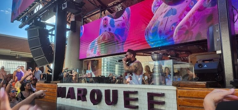 DJ performing at a nightclub with large LED screens displaying colorful visual effects and crowd dancing below the booth