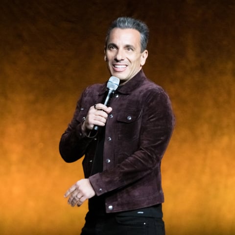 Man smiling while holding a microphone wearing a dark purple velvet jacket against a golden background