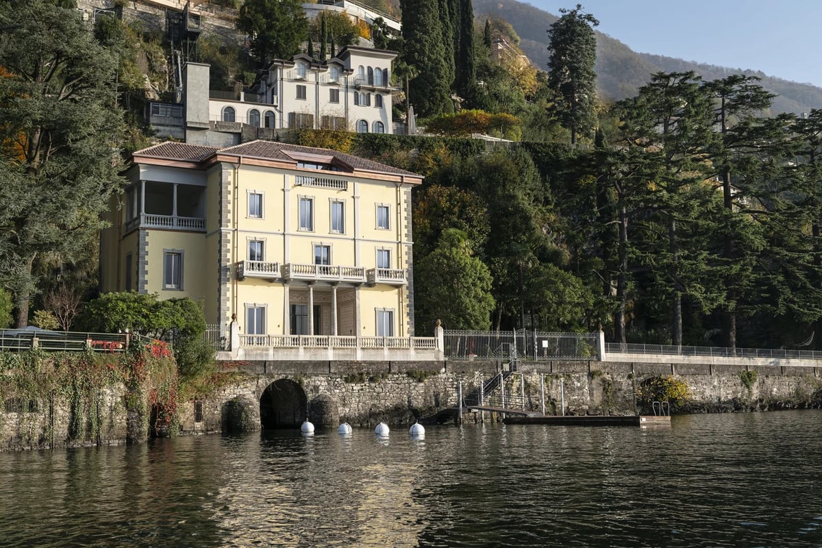 Pale yellow villa with ivy on waterfront of lake, stone wall and archway, mountains and trees in background